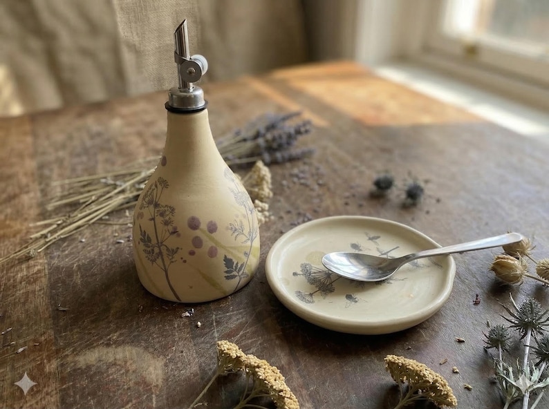 May include: A cream-colored ceramic oil dispenser with a metal pour spout and a matching small round ceramic dish. Both pieces feature a delicate botanical print of dark gray and purple wildflowers. A silver metal spoon rests on the dish.