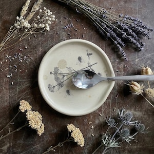 May include: A round, off-white ceramic spoon rest featuring a black botanical illustration of wildflowers and bees. A polished stainless steel spoon rests horizontally across the center of the dish, which has a slightly raised rim and smooth finish.