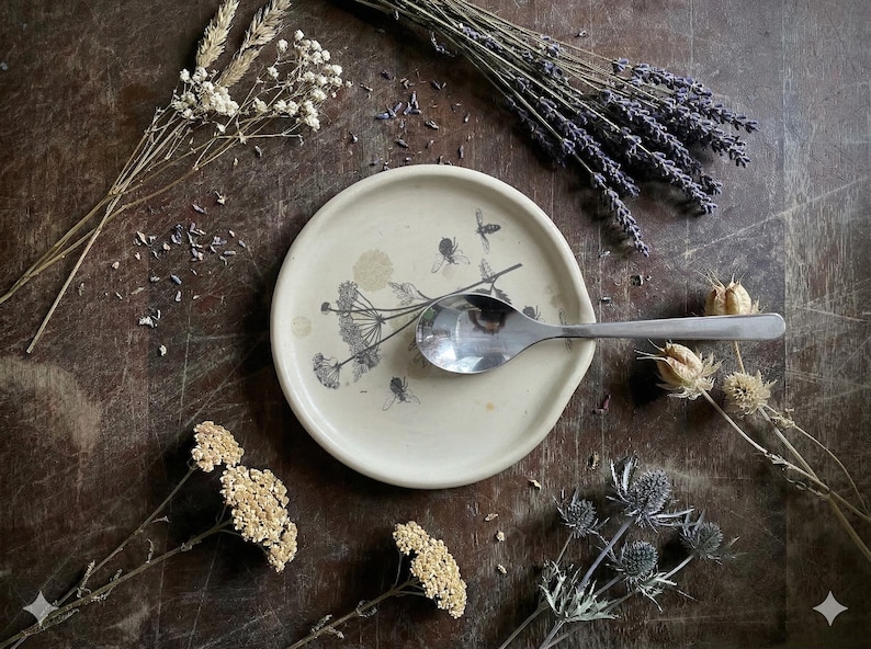 May include: A round, off-white ceramic spoon rest featuring a black botanical illustration of wildflowers and bees. A polished stainless steel spoon rests horizontally across the center of the dish, which has a slightly raised rim and smooth finish.