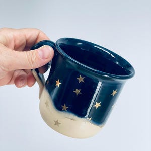 May include: A ceramic mug featuring a two-tone design with a deep navy blue upper section and a cream-colored base. The exterior is decorated with small, metallic gold star motifs scattered across both the blue and cream portions of the mug.