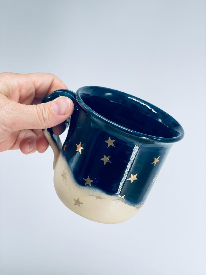 May include: A ceramic mug featuring a two-tone design with a deep navy blue upper section and a cream-colored base. The exterior is decorated with small, metallic gold star motifs scattered across both the blue and cream portions of the mug.