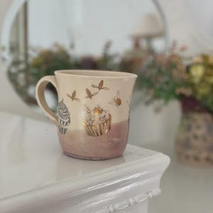 May include: A ceramic mug with a cream-colored upper section and a dusty rose-colored base. The mug features metallic gold illustrations of cupcakes and flying bees encircling the exterior, along with a curved handle on the left side.