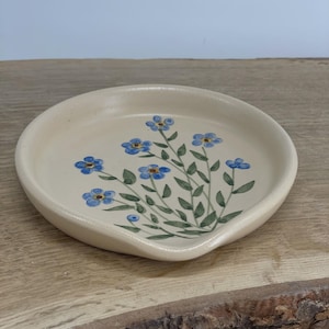 Może przedstawiać: Kremowa ceramiczna podstawka pod łyżkę z kwiatowym wzorem. Podstawka pod łyżkę ma zaokrąglony kształt z małym rantem i malowanym wzorem niebieskich kwiatów i zielonych liści. Podstawka pod łyżkę znajduje się na drewnianej powierzchni.