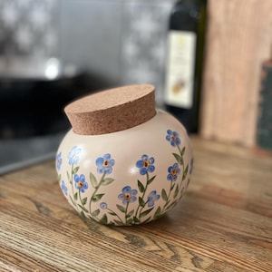 May include: A spherical, cream-coloured ceramic jar with a cork lid, decorated with hand-painted blue forget-me-not flowers and green stems. The jar rests on a wooden surface, displaying a rustic, floral design.