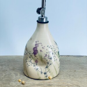 May include: A cream-colored ceramic oil cruet with a rounded base and a tapered neck, featuring a delicate botanical print of dark green stems and purple floral accents. The bottle is topped with a silver-toned metal pourer spout.
