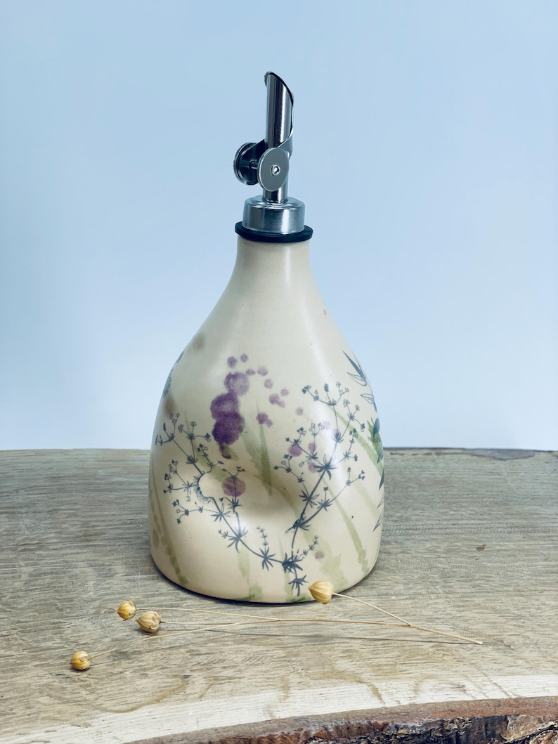 May include: A cream-colored ceramic oil cruet with a rounded base and a tapered neck, featuring a delicate botanical print of dark green stems and purple floral accents. The bottle is topped with a silver-toned metal pourer spout.
