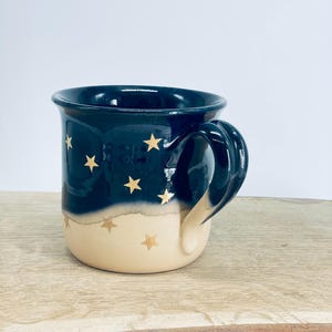 May include: A ceramic mug featuring a two-tone design with a dark navy blue upper section and a cream-colored lower section. The mug is decorated with small, gold-colored star motifs scattered across both the dark and light portions of the exterior.