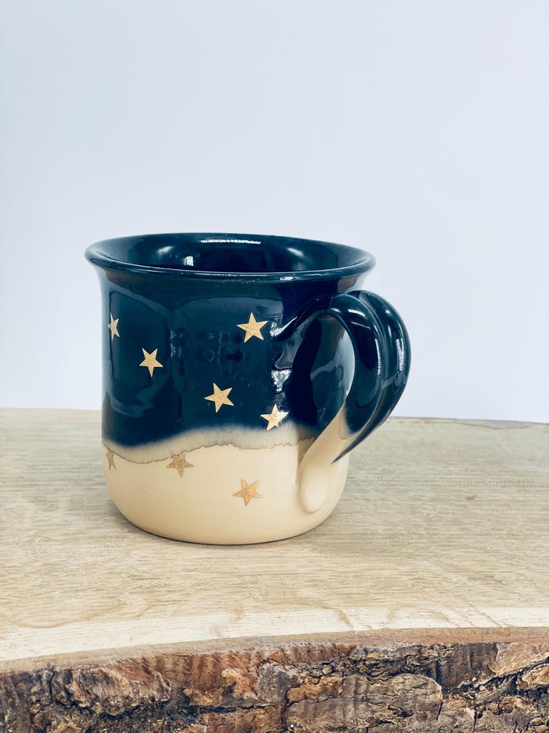 May include: A ceramic mug featuring a two-tone design with a dark navy blue upper section and a cream-colored lower section. The mug is decorated with small, gold-colored star motifs scattered across both the dark and light portions of the exterior.