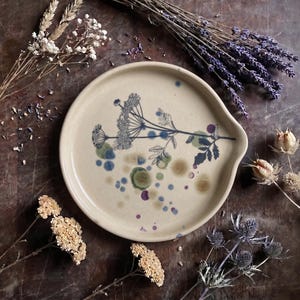 Könnte beinhalten: Eine runde, cremefarbene Keramik-Löffelablage mit einer dunkelblauen botanischen Illustration einer blühenden Pflanze. Die Oberfläche ist mit verstreuten, durchscheinenden blauen, grünen und violetten kreisförmigen Punkten um das zentrale Motiv verziert.