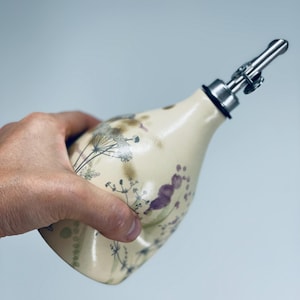 May include: A cream-colored ceramic oil dispenser with a tapered neck and a stainless steel pour spout. The body features a delicate botanical print of dark purple and black wildflowers, stems, and seed heads scattered across the smooth surface.
