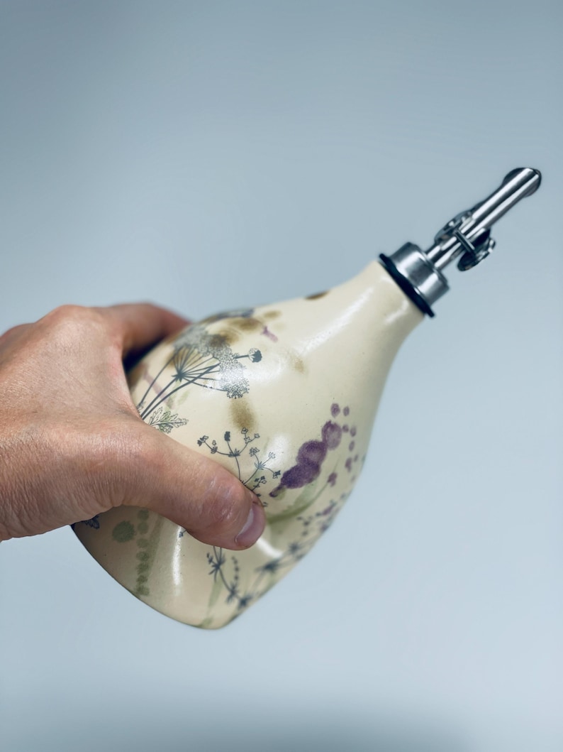 May include: A cream-colored ceramic oil dispenser with a tapered neck and a stainless steel pour spout. The body features a delicate botanical print of dark purple and black wildflowers, stems, and seed heads scattered across the smooth surface.