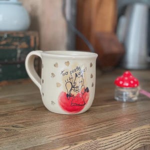 May include: A cream-colored ceramic mug featuring a hand-drawn illustration of a fox in a red dress, a red circular accent, and gold heart motifs. The text "Too pretty to wake!" is written in black ink above the fox illustration on the mug.
