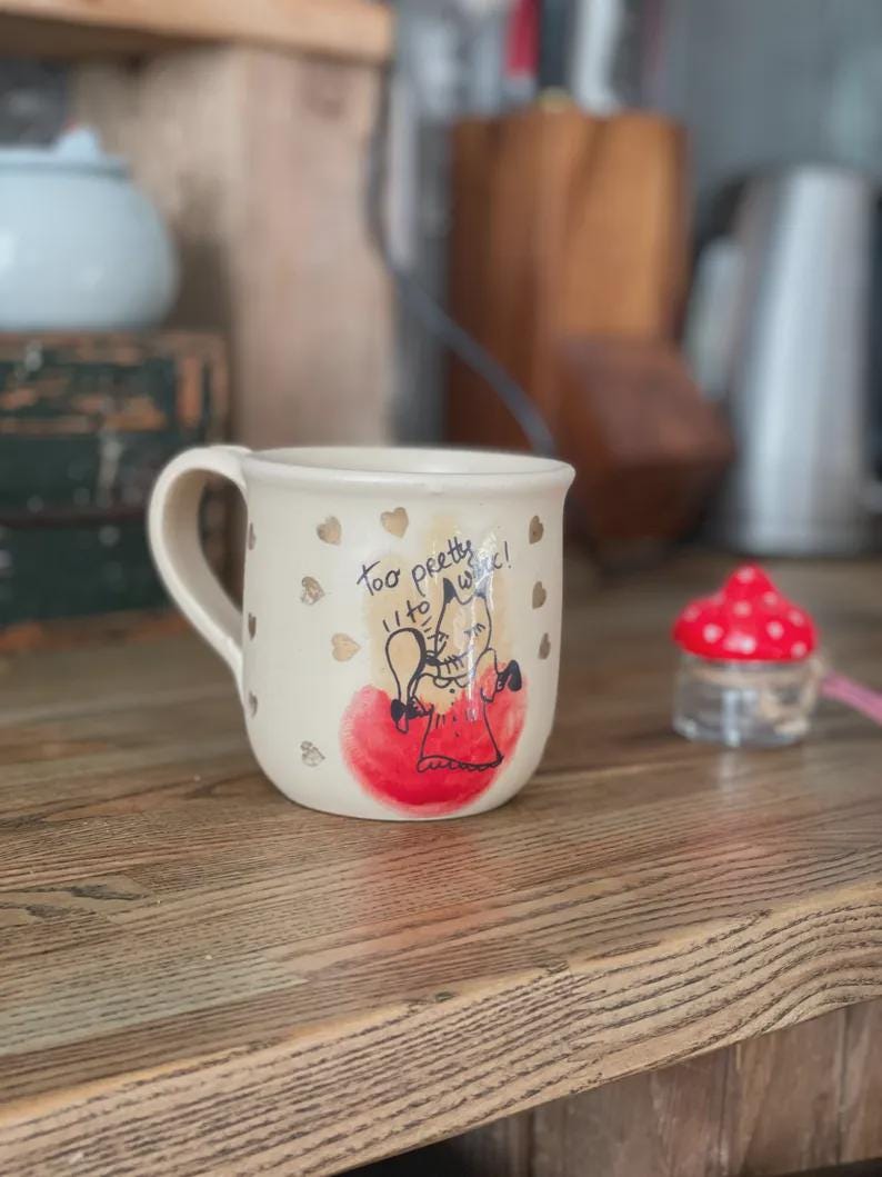 May include: A cream-colored ceramic mug featuring a hand-drawn illustration of a fox in a red dress, a red circular accent, and gold heart motifs. The text "Too pretty to wake!" is written in black ink above the fox illustration on the mug.