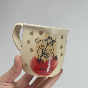 May include: A cream-colored ceramic mug featuring a hand-drawn illustration of a fox holding a mirror, with the text "too pretty to work!" written above it. The mug is decorated with small gold hearts and a red watercolor-style circular accent.