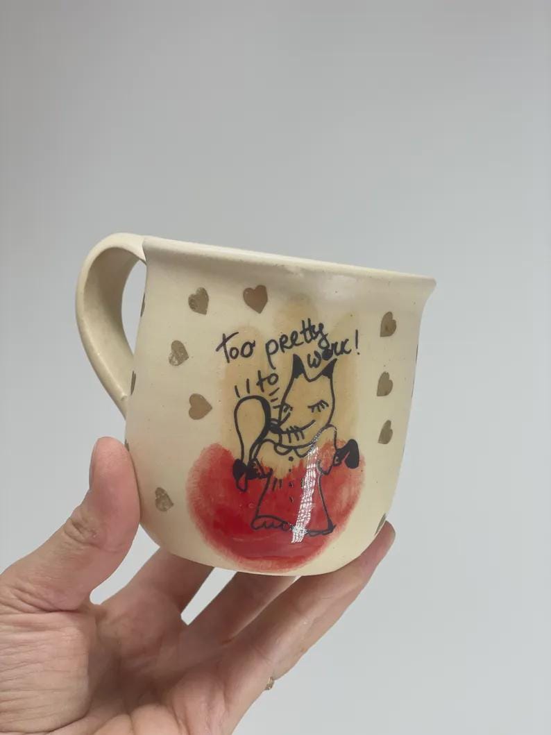 May include: A cream-colored ceramic mug featuring a hand-drawn illustration of a fox holding a mirror, with the text "too pretty to work!" written above it. The mug is decorated with small gold hearts and a red watercolor-style circular accent.