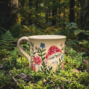 Peut inclure: Mug en céramique crème orné de champignons rouges et de fleurs bleues. Le mug est posé dans un décor forestier avec de la mousse et du feuillage verts. Le design du mug comprend des champignons et des éléments floraux peints à la main.