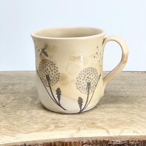 May include: Cream ceramic mug with a dandelion design. The mug has a curved handle and a flared rim. The dandelion design is in dark grey, with a dragonfly and other small details. The mug is on a wooden surface.
