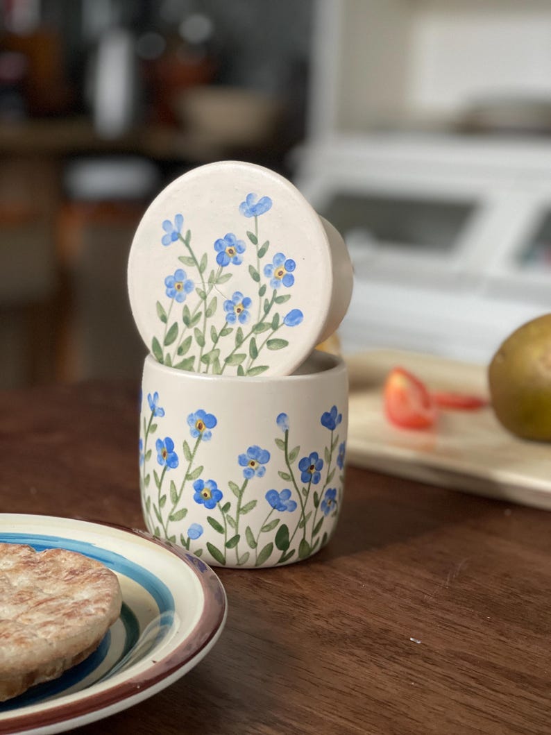 May include: A white ceramic butter dish with a lid, both decorated with blue forget-me-not flowers. The dish is sitting on a wooden table.