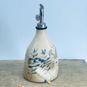May include: A cream-colored ceramic oil dispenser with a tapered body and a metallic pour spout. The surface features dark gray botanical line drawings of leaves and stems, accented by scattered blue paint splatters and soft green washes.