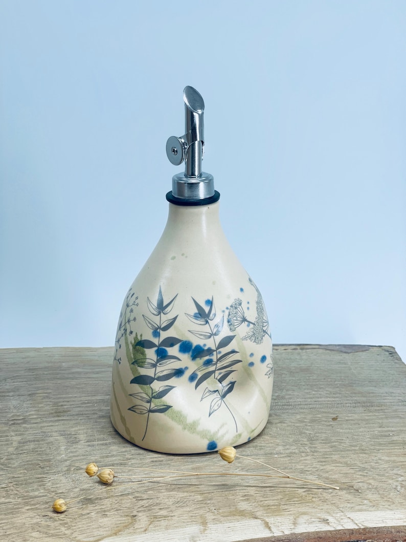 May include: A cream-colored ceramic oil dispenser with a tapered body and a metallic pour spout. The surface features dark gray botanical line drawings of leaves and stems, accented by scattered blue paint splatters and soft green washes.