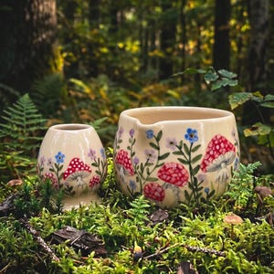 May include: A cream-colored ceramic creamer and a matching small egg-shaped vessel, both hand-painted with red-capped mushrooms, green stems, and small blue and purple flowers. The items feature a smooth, glossy finish and rounded forms.