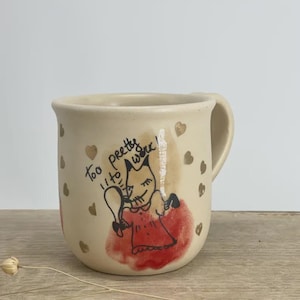 May include: A beige ceramic mug featuring a hand-drawn illustration of a fox character in a red dress holding a mirror. The text "too pretty to work!" is written above the fox, surrounded by small gold heart accents scattered across the mug surface.