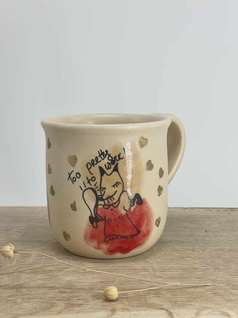 May include: A beige ceramic mug featuring a hand-drawn illustration of a fox character in a red dress holding a mirror. The text "too pretty to work!" is written above the fox, surrounded by small gold heart accents scattered across the mug surface.