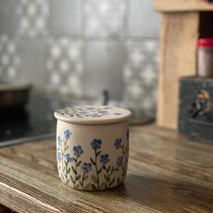 May include: A white ceramic jar with a lid, decorated with blue forget-me-not flowers and green stems. The jar is sitting on a wooden surface.