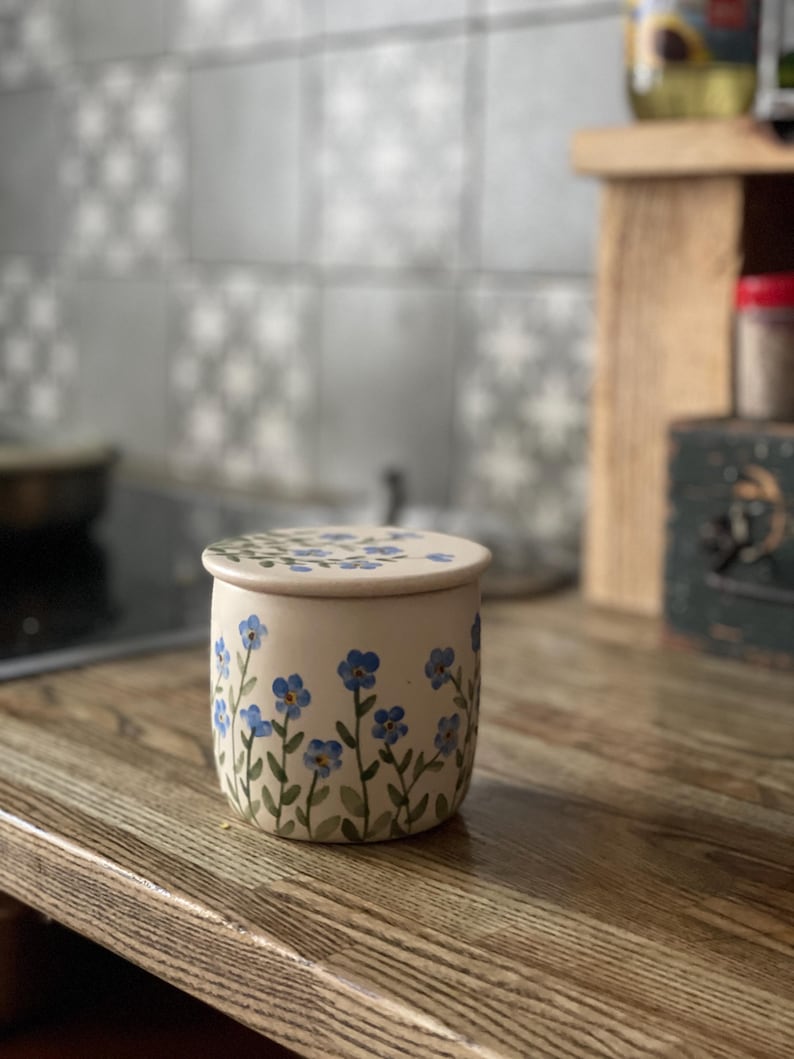 May include: A white ceramic jar with a lid, decorated with blue forget-me-not flowers and green stems. The jar is sitting on a wooden surface.