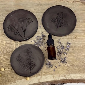 Könnte beinhalten: Drei kreisförmige, dunkelgraue Tonteller mit geprägten botanischen Motiven von Blumen und Zweigen. Eine kleine braune Glastropfflasche mit schwarzem Verschluss steht in der Mitte, umgeben von verstreuten getrockneten Lavendelblüten.