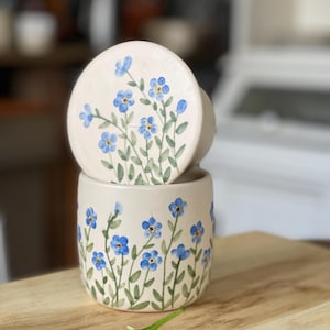 May include: A ceramic container with a lid, both decorated with hand-painted blue forget-me-not flowers on a white background. The container is sitting on a wooden surface.