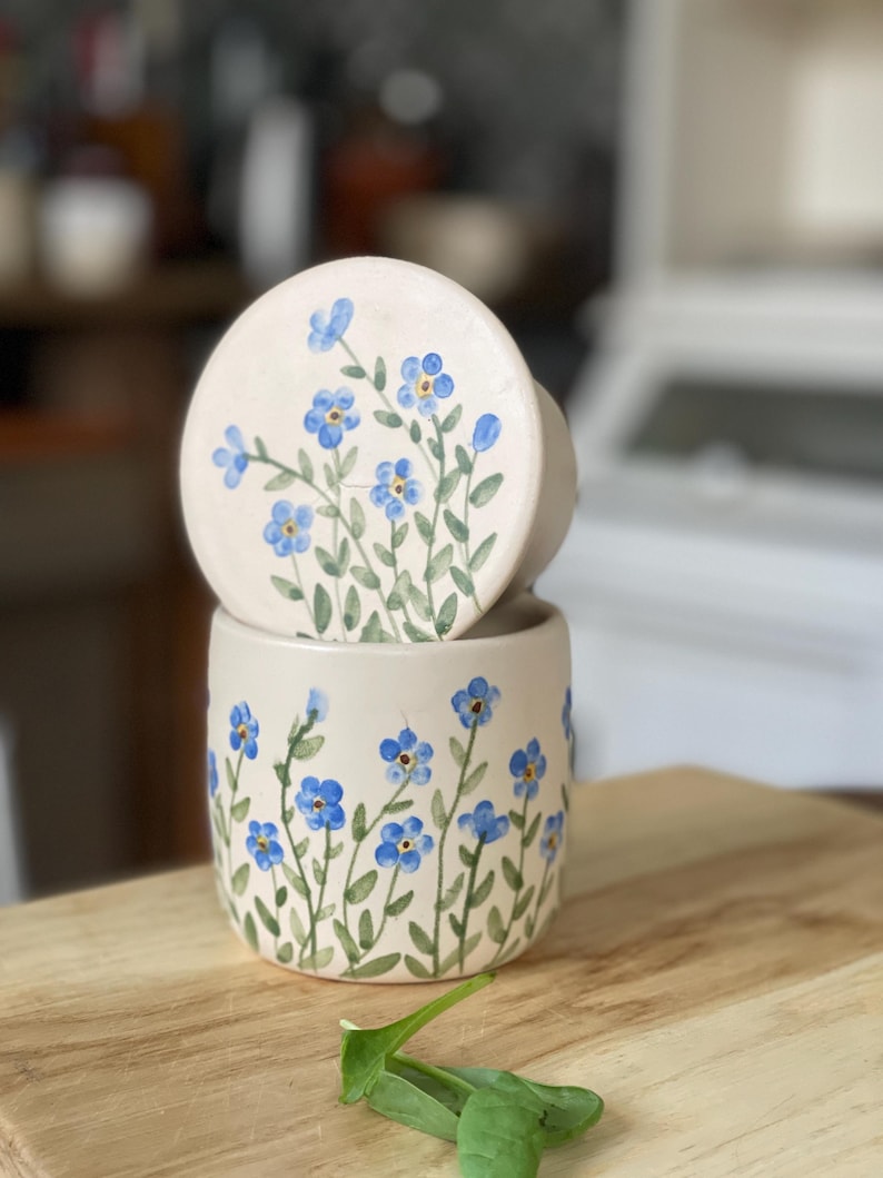 May include: A ceramic container with a lid, both decorated with hand-painted blue forget-me-not flowers on a white background. The container is sitting on a wooden surface.