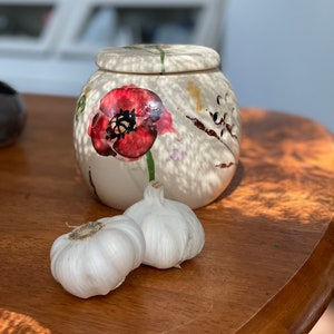 Può includere: Un vaso di ceramica bianca con un disegno floreale con un papavero rosso. Due spicchi d'aglio bianchi sono davanti al vaso su un tavolo di legno.