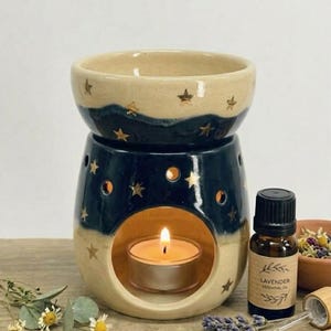 May include: A ceramic oil burner with a two-tone cream and dark blue glaze featuring gold star accents. The burner has a circular opening containing a lit tea light candle. Beside it sits a small brown bottle labelled Lavender Essential Oil.