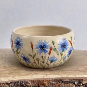 Op de afbeelding: Een ronde, crèmekleurige keramische kom met een handgeschilderd botanisch ontwerp. De buitenkant is versierd met levendige blauwe korenbloemen, groene stengels, bladeren en dunne, roodbruine stengels in een herhalend bloemenpatroon.