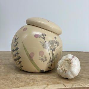 Puede incluir: Un tarro de cerámica con tapa, decorado con un patrón floral en tonos verdes, marrones y rosas. El tarro está sentado sobre una superficie de madera junto a un solo diente de ajo.