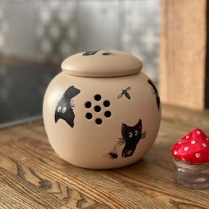 Puede incluir: Un tarro de cerámica beige con tapa, decorado con gatos negros y moscas. El tarro tiene pequeños agujeros en el lateral y está sobre una superficie de madera.