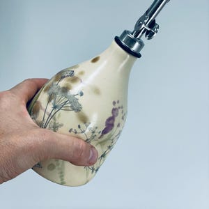 May include: A cream-colored ceramic oil dispenser with a tapered neck and a stainless steel pour spout. The body features a botanical print of dark green and purple wildflowers and grasses, accented by soft brown smudges across the surface.