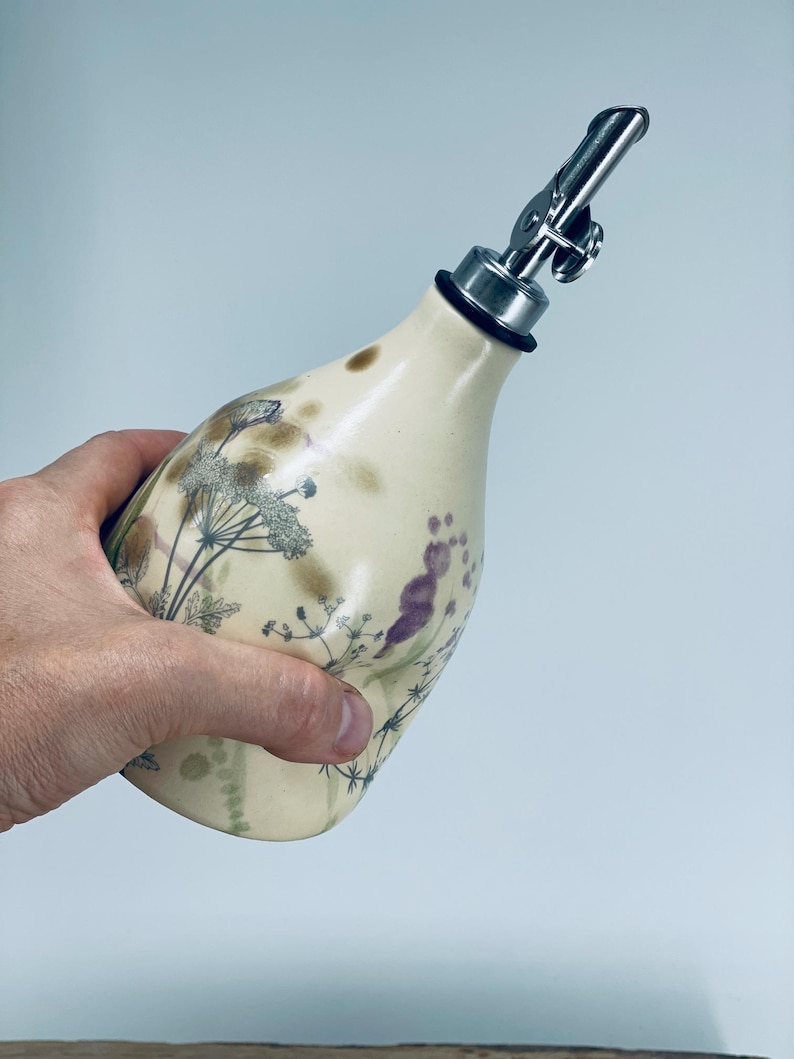 May include: A cream-colored ceramic oil dispenser with a tapered neck and a stainless steel pour spout. The body features a botanical print of dark green and purple wildflowers and grasses, accented by soft brown smudges across the surface.
