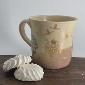 May include: A ceramic mug with a two-tone cream and dusty rose glaze, featuring gold-colored illustrations of cupcakes and bees. Two white, swirled meringue cookies are positioned in front of the mug, which has a curved handle.