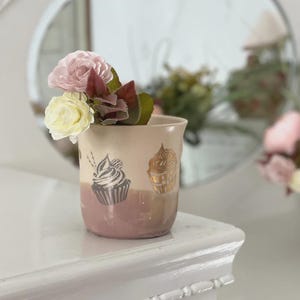 May include: A ceramic cup featuring a two-tone design with a cream upper section and a mauve lower section. The cup is decorated with two cupcake illustrations, one in dark grey and one in gold, and contains two artificial flowers in pink and cream.