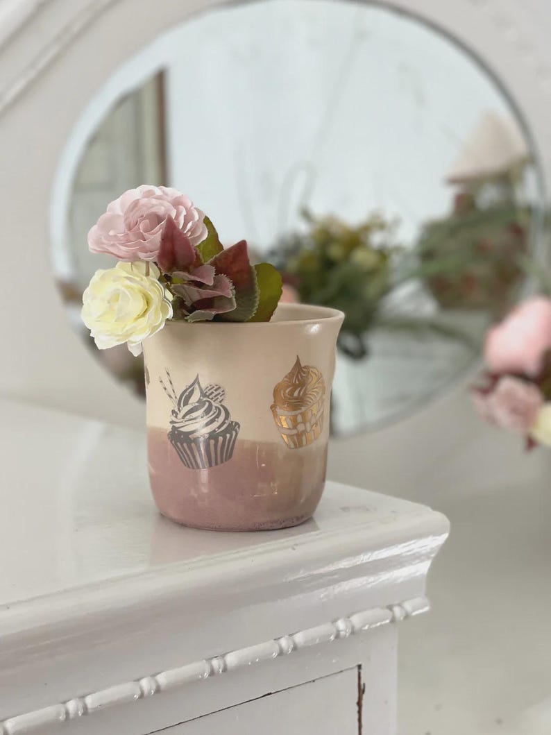 May include: A ceramic cup featuring a two-tone design with a cream upper section and a mauve lower section. The cup is decorated with two cupcake illustrations, one in dark grey and one in gold, and contains two artificial flowers in pink and cream.
