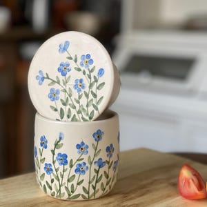 May include: A white ceramic jar with a lid, both decorated with hand-painted blue forget-me-not flowers on green stems. The jar is sitting on a wooden surface.