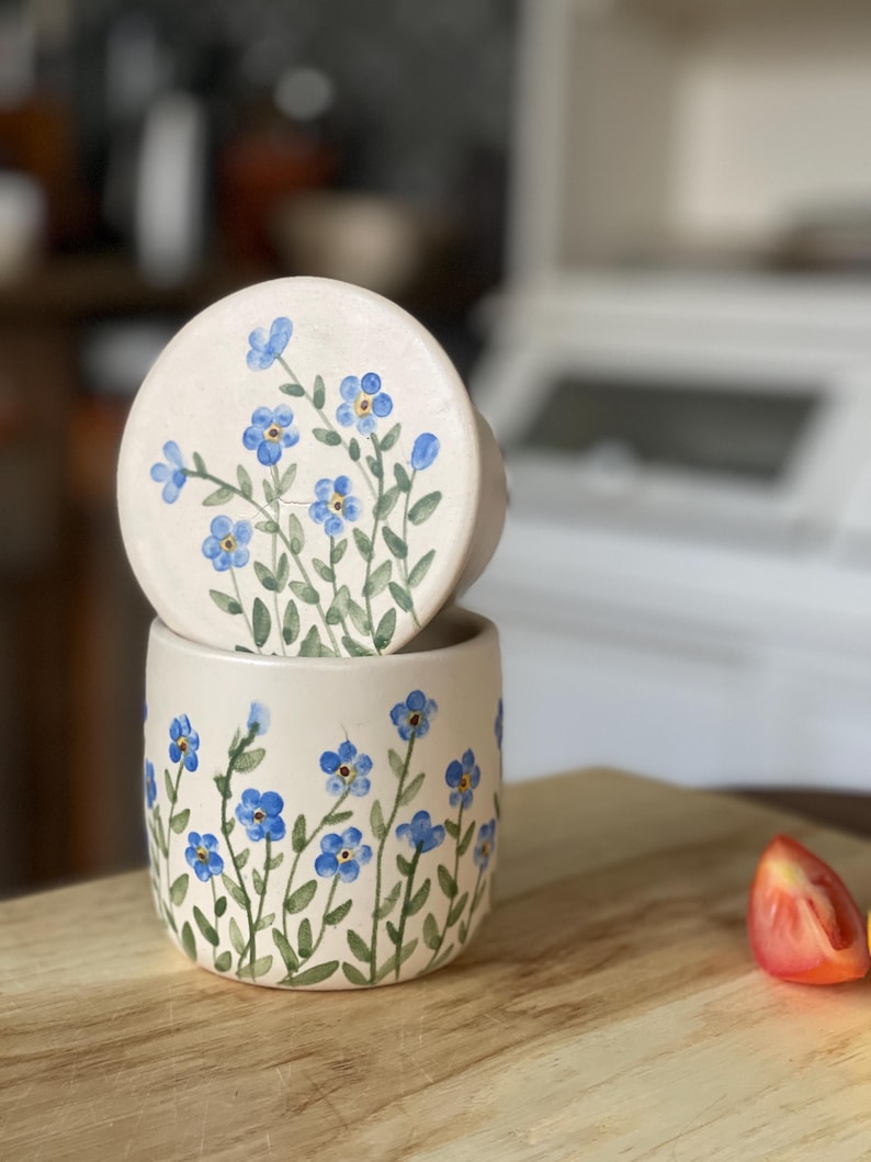 May include: A white ceramic jar with a lid, both decorated with hand-painted blue forget-me-not flowers on green stems. The jar is sitting on a wooden surface.