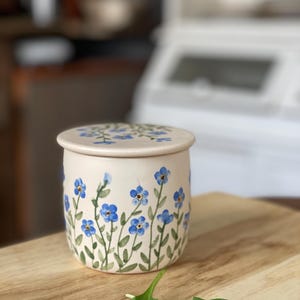 May include: A ceramic butter dish with a lid, featuring a floral design of blue forget-me-not flowers and green stems on a cream background.