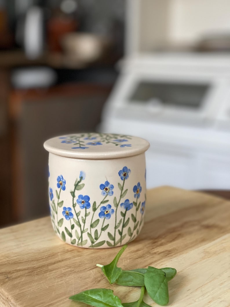 May include: A ceramic butter dish with a lid, featuring a floral design of blue forget-me-not flowers and green stems on a cream background.