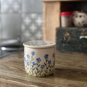 May include: A white ceramic butter dish with a lid, decorated with blue forget-me-not flowers and green stems. The dish is sitting on a wooden surface.