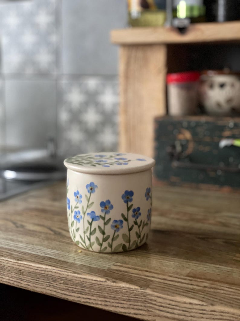 May include: A white ceramic butter dish with a lid, decorated with blue forget-me-not flowers and green stems. The dish is sitting on a wooden surface.