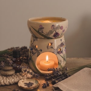 Peut inclure: Un brûle-parfum en céramique avec une base crème et un bol supérieur, orné d'un motif botanique de fleurs sauvages violettes et vertes. Une bougie chauffe-plat allumée est placée dans l'ouverture circulaire et le bol contient de l'huile.