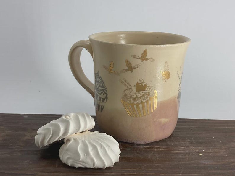 May include: A ceramic mug with a two-tone cream and dusty rose glaze, featuring gold-colored illustrations of cupcakes and bees. Two white, swirled meringue cookies are positioned in front of the mug, which has a curved handle.